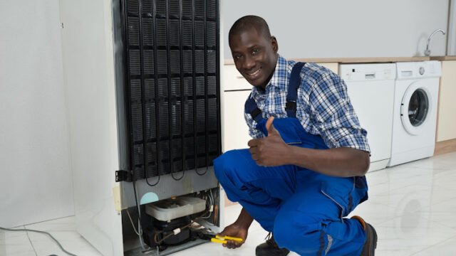 Young Happy African Technician In Uniform Fixing Refrigerator In Kitchen Room