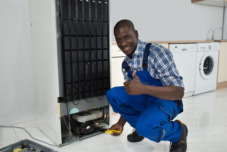 Young Happy African Technician In Uniform Fixing Refrigerator In Kitchen Room